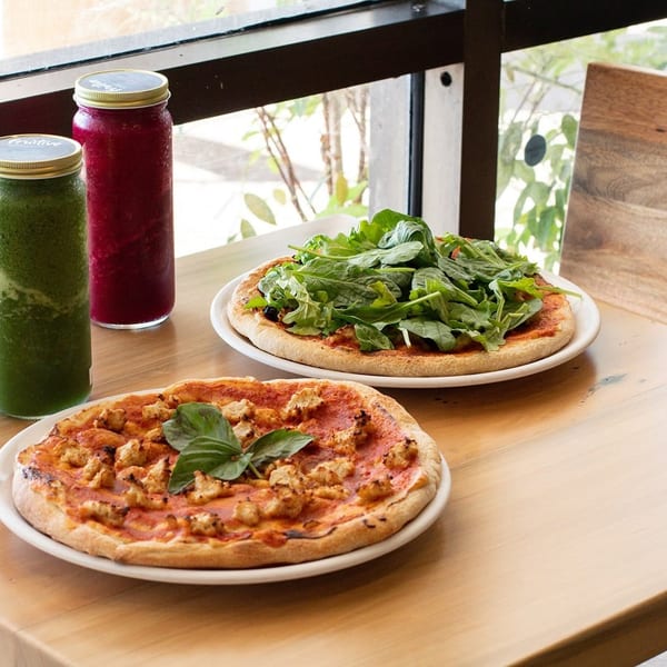 Two pizzas, one with chicken and basil, the other with a pile of greens, next to two smoothies.
