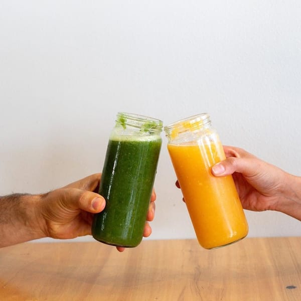 Two hands clinking glass bottles of green and orange smoothies over a wooden table.
