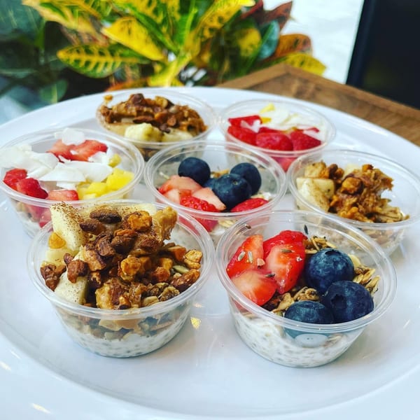 Seven small clear cups of yogurt parfaits with various fruit and granola toppings on a white plate.