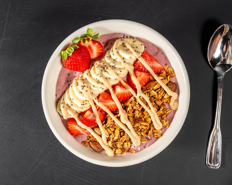 Overhead view of a smoothie bowl with strawberries, bananas, granola, chia seeds, and peanut butter drizzle.