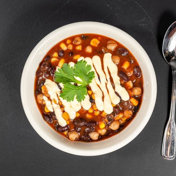 Overhead view of a bowl of chili with black beans, corn, and chickpeas, garnished with cilantro and white sauce, next to a spoon.