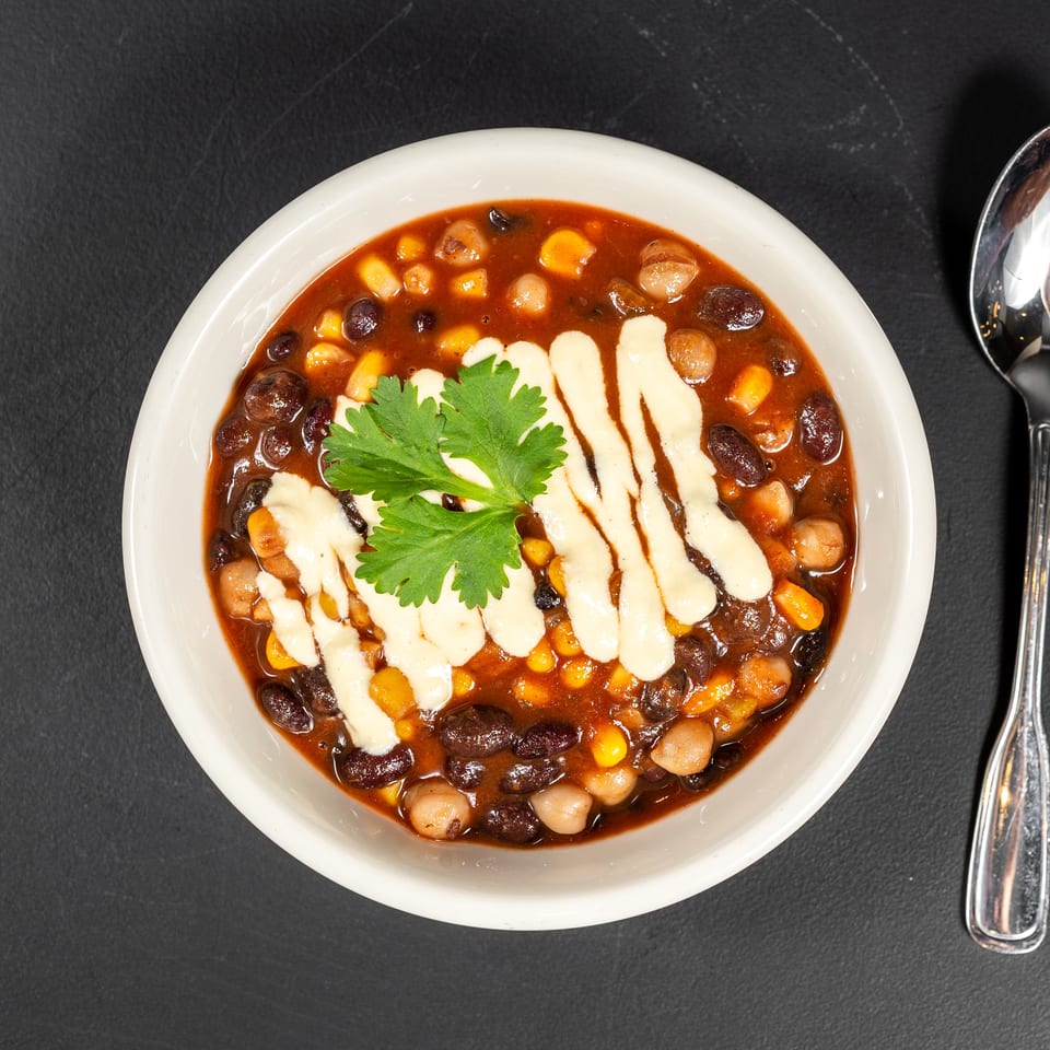 Overhead view of a bowl of chili with black beans, corn, and chickpeas, garnished with cilantro and white sauce, next to a spoon.