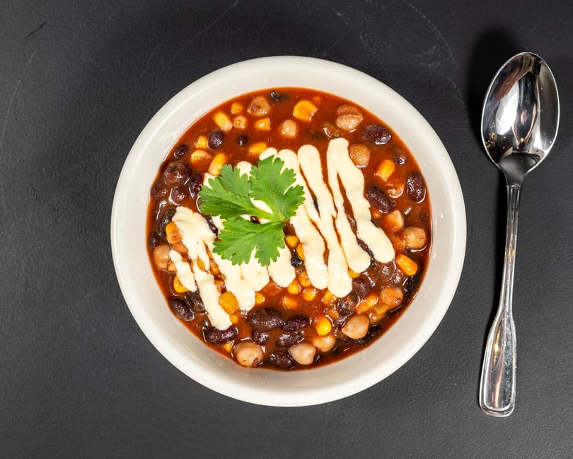 Overhead view of a bowl of chili with black beans, corn, and chickpeas, garnished with cilantro and white sauce, next to a spoon.