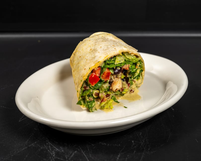A colorful wrap filled with lettuce, tomatoes, olives, and a creamy dressing on a white plate.