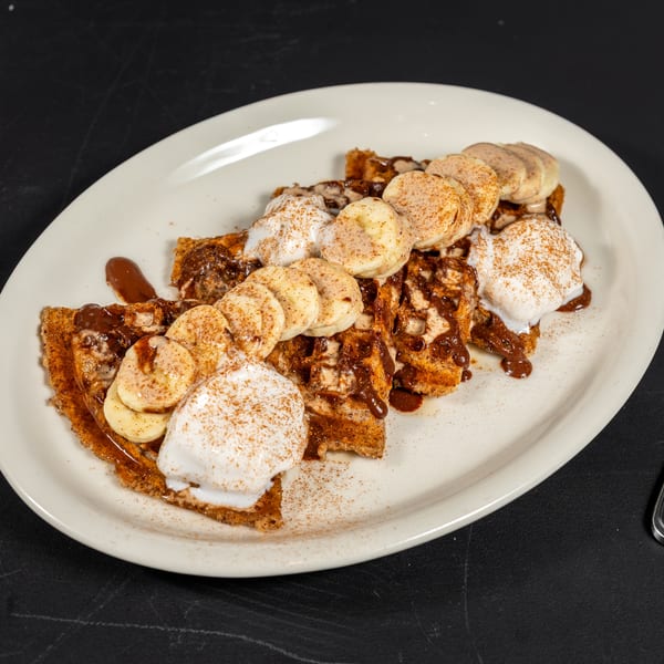 Waffles topped with sliced bananas, whipped cream, chocolate syrup, and cinnamon on a white plate.