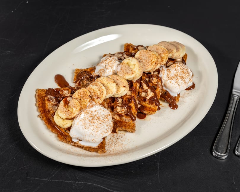 Waffles topped with sliced bananas, whipped cream, chocolate syrup, and cinnamon on a white plate.