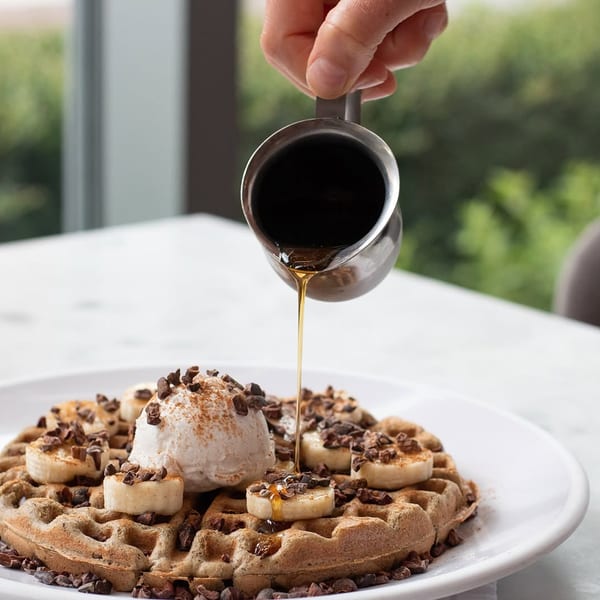 Maple syrup being poured over a waffle with bananas, ice cream, and chocolate nibs.