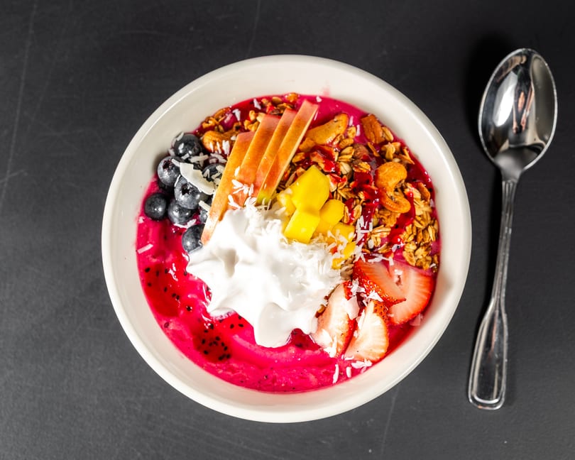 Vibrant pink smoothie bowl topped with blueberries, sliced apples, mango, strawberries, granola, and coconut flakes.